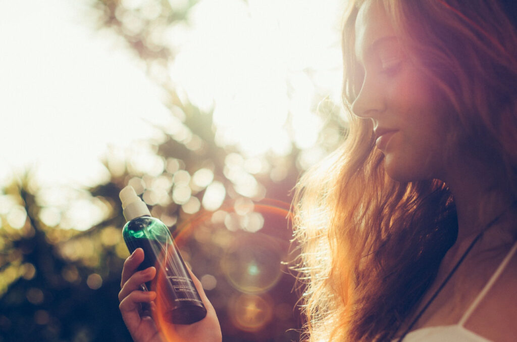 A woman with red hair holding a green skincare bottle with the sun reflecting from behind her.